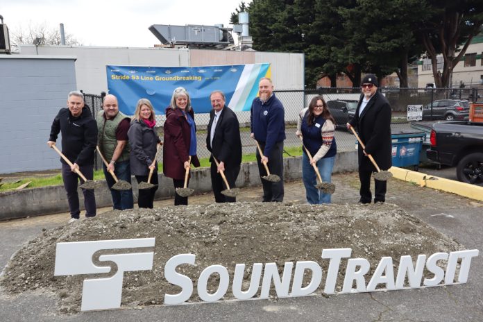 Eight Sound Transit leaders lift a shovelful of ceremonial dirt next to an agency logo and poster board