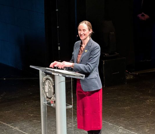 Wilson Sketches Affordability Agenda in State of City Speech Wilson smiles and stands at a lectern with the City seal. She wears a red skirt and gray blazer.