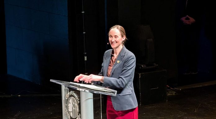 Wilson Sketches Affordability Agenda in State of City Speech Wilson smiles and stands at a lectern with the City seal. She wears a red skirt and gray blazer.