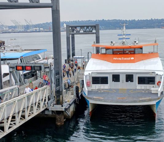 Mosquito Fleet Act Runs Into Choppy Seas in the State Senate Passengers load onto a orange and white Kitsap fast ferry boat with Elliott Bay in the background.