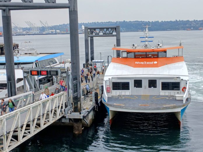 Passengers load onto a orange and white Kitsap fast ferry boat with Elliott Bay in the background.