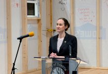 Wilson Rolls Out Bills Expediting Emergency Housing for Homeless Residents Wilson wears a black blazer and stands at a lectern in front of a half completed tiny home with inspirational messages scrawled on the siding.