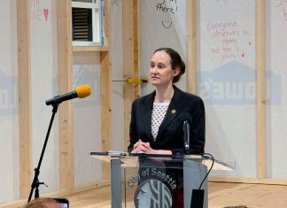 Wilson Rolls Out Bills Expediting Emergency Housing for Homeless Residents Wilson wears a black blazer and stands at a lectern in front of a half completed tiny home with inspirational messages scrawled on the siding.