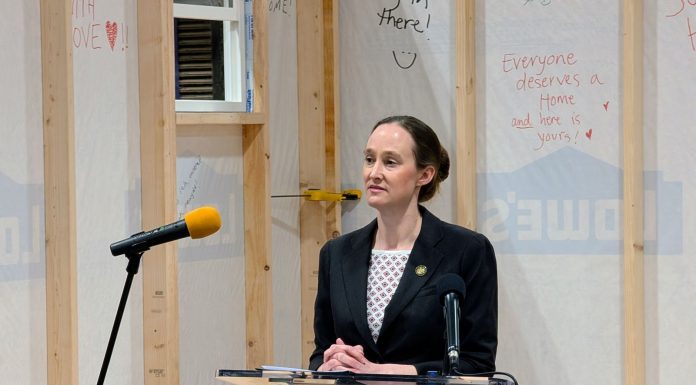 Wilson Rolls Out Bills Expediting Emergency Housing for Homeless Residents Wilson wears a black blazer and stands at a lectern in front of a half completed tiny home with inspirational messages scrawled on the siding.